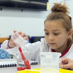 A young girl in a lab coat pouring a red liquid into a beaker