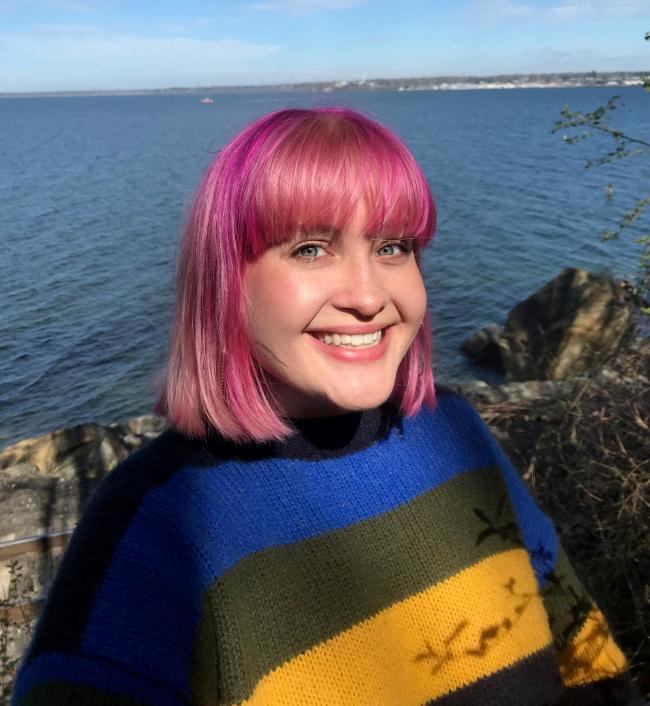 Shannon, with pink hair and wearing a boldy striped sweater, posing in front of a rocky shoreline